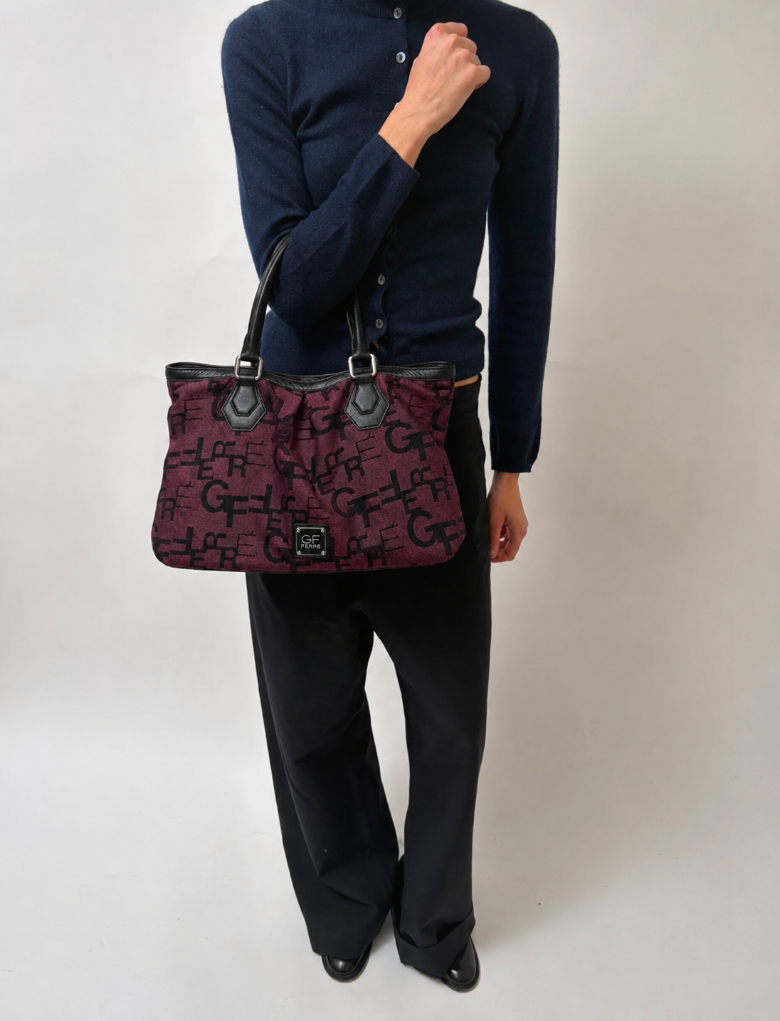 Person wearing a dark sweater and black pants holding a patterned handbag against a plain background